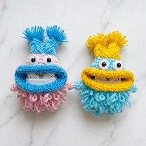 May include: Two handmade crochet AirPod cases in the shape of whimsical monsters. One is pink and blue, the other is blue and yellow. Each has a wide open mouth, two eyes, and a fluffy body. The cases are designed to hold wireless earbuds.