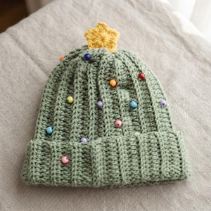 May include: A sage green crocheted beanie with a ribbed texture, featuring a yellow crocheted star at the crown and scattered multicolored plastic beads throughout the body of the hat, resembling a decorated evergreen tree.