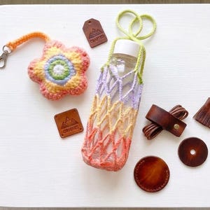 May include: A collection of handmade items, including a crocheted flower keychain, a gradient-colored bottle holder, and leather accessories. The bottle holder is designed to fit a clear glass bottle. The color palette includes orange, yellow, purple, and green.
