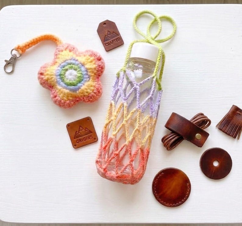 May include: A collection of handmade items, including a crocheted flower keychain, a gradient-colored bottle holder, and leather accessories. The bottle holder is designed to fit a clear glass bottle. The color palette includes orange, yellow, purple, and green.