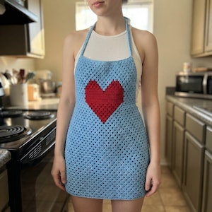 May include: A light blue crochet apron featuring a prominent red heart design in the center. The apron is constructed with a classic granny square stitch pattern and includes a halter-style neck strap that secures behind the neck.