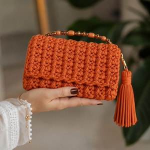 Puede incluir: Un bolso de mano rectangular de ganchillo naranja con un patrón de puntos texturizado, un asa superior adornada con cuentas naranjas y metálicas, y una borla de tela naranja larga y gruesa sujeta al lateral con un cierre de metal dorado.
