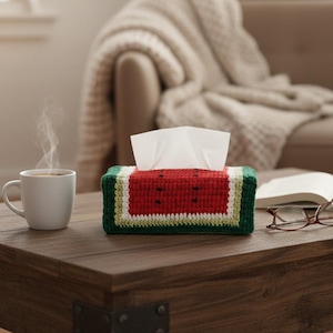 Może przedstawiać: Ręcznie robiona osłona na pudełko chusteczek w kształcie arbuza. Pokrywa jest szydełkowana czerwoną, zieloną i białą przędzą, z czarnymi detalami nasion. Widoczne są białe chusteczki. Biały kubek kawy i książka z okularami stoją na drewnianym stole.