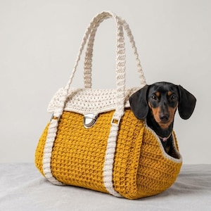 May include: A mustard yellow and cream crochet dog carrier bag with a black and brown dachshund dog inside. The bag has a flap closure and cream-colored handles and trim. The dog is looking at the camera.