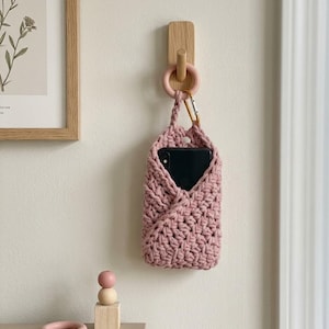 Puede incluir: Un soporte para teléfono de ganchillo rosa polvoriento con un mosquetón dorado, colgado de una clavija de madera en una pared color crema. Un teléfono inteligente negro está dentro. También se ven una impresión botánica enmarcada y juguetes de madera.