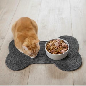 May include: A ginger cat eating from a white bowl of food on a gray, bone-shaped mat. The mat is made of a woven material and sits on a light wood floor. The cat is centered in the image.
