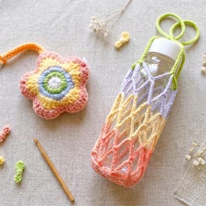 May include: A crocheted flower keychain with a silver clasp and a gradient water bottle holder. The holder is designed to fit a clear bottle. The flower keychain and bottle holder are in shades of pink, yellow, and green.