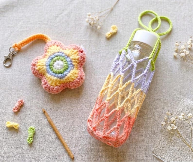 May include: A crocheted flower keychain with a silver clasp and a gradient water bottle holder. The holder is designed to fit a clear bottle. The flower keychain and bottle holder are in shades of pink, yellow, and green.