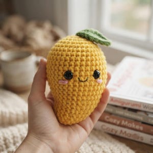 May include: A yellow crochet mango amigurumi with a green leaf on top. The fruit features two black plastic safety eyes, a small embroidered smile, and two pink stitched cheeks. The texture is created with tight yarn stitches in a rounded shape.