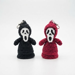 May include: Two handmade crochet keychains shaped like hooded figures with white ghost-like masks featuring black eye and mouth cutouts. One figure is crafted in solid black yarn, while the other is in deep red yarn, both with metal key rings attached.