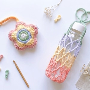 May include: A crocheted flower-shaped keychain and a water bottle holder. The holder is a net-like design in pastel colors, and the keychain has a flower design with a silver clasp. A wooden crochet hook and small yarn pieces are also present.