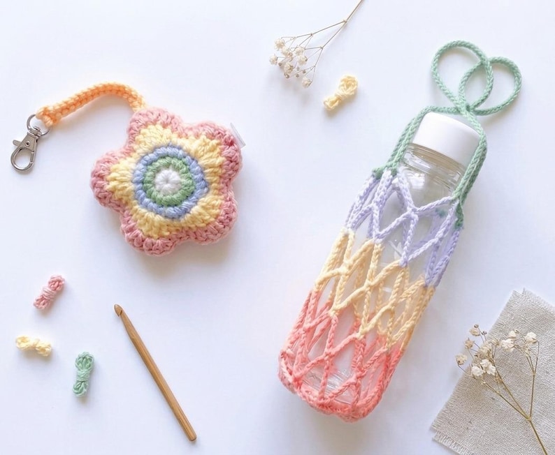 May include: A crocheted flower-shaped keychain and a water bottle holder. The holder is a net-like design in pastel colors, and the keychain has a flower design with a silver clasp. A wooden crochet hook and small yarn pieces are also present.