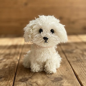 May include: A small, white, plush dog figurine crafted with a textured, shaggy yarn finish. The toy features two round, black, glossy eyes, a small black stitched nose, and a light pink collar around its neck, sitting in an upright position.