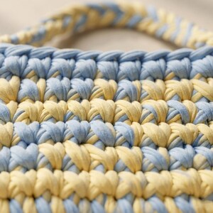 May include: Close-up of a handmade crochet bag in a repeating pattern of light blue and yellow. The bag has a matching handle. The texture of the yarn is visible, creating a soft, textured appearance. The bag is likely used for carrying items.