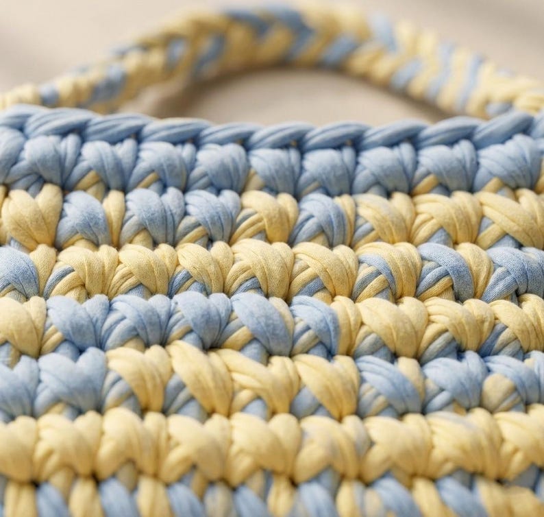 May include: Close-up of a handmade crochet bag in a repeating pattern of light blue and yellow. The bag has a matching handle. The texture of the yarn is visible, creating a soft, textured appearance. The bag is likely used for carrying items.