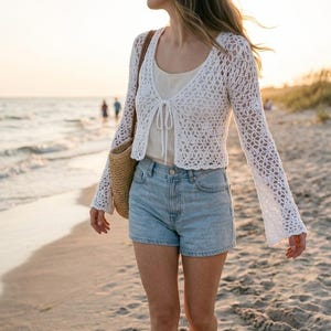 May include: A white, open-weave crochet cardigan with long, flared sleeves. The cardigan is paired with a light-colored top and light blue denim shorts. A woven straw bag is carried over the shoulder. The scene is set on a beach at sunset.