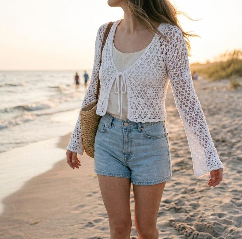 May include: A white, open-weave crochet cardigan with long, flared sleeves. The cardigan is paired with a light-colored top and light blue denim shorts. A woven straw bag is carried over the shoulder. The scene is set on a beach at sunset.
