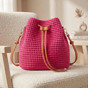 May include: A vibrant fuchsia pink crochet bucket bag featuring a textured knit pattern, a tan leather drawstring closure, and matching tan leather shoulder straps attached with gold-toned metal hardware clips on both sides of the bag.