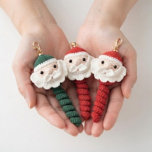 May include: Three crochet Santa Claus keychains with gold-tone lobster clasps. Each features a round head with a white beard, black bead eyes, and a spiral-shaped body. One Santa has a green hat and body, while the other two have red hats and bodies.