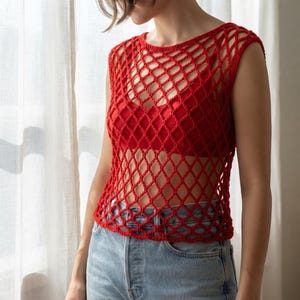 May include: A bright red sleeveless crochet top featuring an open diamond-shaped mesh pattern. The garment is worn over a solid red base layer and is paired with light blue denim jeans, creating a layered look with a distinct textured finish.