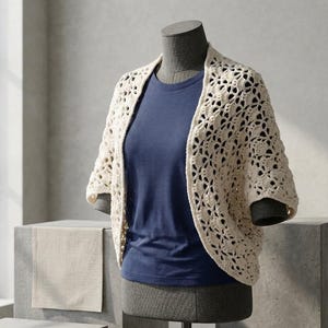 Op de afbeelding: Een crèmekleurige, open geweven gehaakte omslagdoek met korte mouwen wordt getoond op een mannequin. De omslagdoek wordt gedragen over een marineblauw t-shirt. Het open ontwerp van het haakwerk creëert een delicate, luchtige uitstraling. De mannequin staat op een grijs voetstuk.