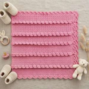 May include: A square pink crochet blanket with a scalloped edge, accompanied by two pairs of cream-coloured knit baby booties, a wooden teething ring with bunny ears, a pink dummy, and a small cream-coloured plush teddy bear toy.
