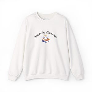 May include: A white long-sleeve crewneck sweatshirt featuring a small graphic centered on the chest. The graphic depicts a stack of three books with a small yellow flower on top, accompanied by the black text Cursed by Literature in an arc.