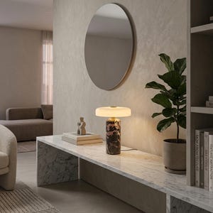 May include: A modern interior scene featuring a marble-topped console table with a decorative lamp. The lamp has a cylindrical base with a marble pattern and a round, white shade. A round mirror hangs on the wall above the table.