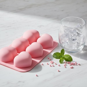 Peut inclure: Un bac à glaçons en silicone rose avec des moules en forme de cœur. Un verre transparent rempli de glaçons est posé à côté du bac. Des feuilles de menthe fraîche et des vermicelles roses sont dispersés.