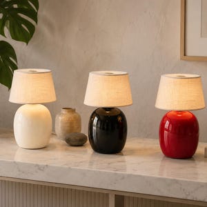 May include: Three table lamps with rounded, glossy ceramic bases in white, black, and red. Each lamp features a tapered, light-colored fabric drum shade. The lamps are arranged in a row, emitting a soft, warm glow from within the shades.