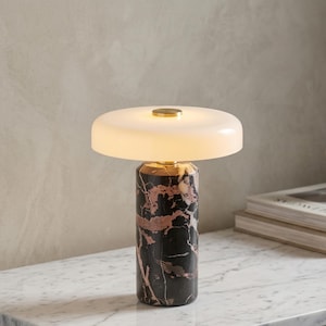 Puede incluir: Una lámpara de mesa moderna con una base cilíndrica de mármol negro con vetas rosas. La lámpara tiene una pantalla blanca en forma de disco y un acento dorado. La lámpara está sobre una superficie de mármol.