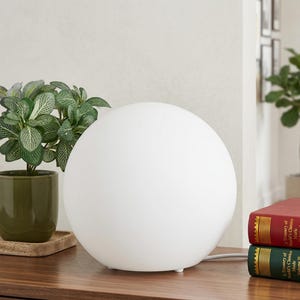 Op de afbeelding: Een witte, bolvormige tafellamp met een glad oppervlak. De lamp staat op een houten oppervlak, naast een kleine potplant en twee boeken. Het ontwerp van de lamp is minimalistisch en modern.
