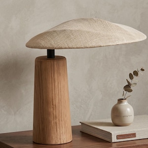 Könnte beinhalten: Eine moderne Tischlampe mit einem breiten, geschwungenen, hellbeigen Schirm und einem zylindrischen Holzsockel. Die Lampe steht auf einer Holzoberfläche, neben einer kleinen Vase mit einem Zweig Grün.
