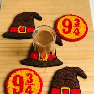 May include: A set of four handmade coasters. Two are shaped like brown wizard hats with red bands and gold buckles. The other two are yellow circles with red trim and the number 9 3/4 in red. A glass of coffee sits on one of the hat coasters.