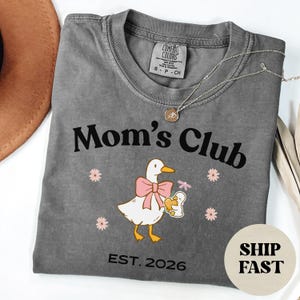 May include: A grey Comfort Colors t-shirt with the text "Mom's Club" and a graphic of a white duck wearing a pink bow, holding a baby duck. The shirt also features the text "EST. 2026" and small pink flowers.