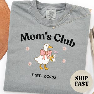 May include: A grey t-shirt with the words "Mom's Club" in black. The shirt features a cartoon duck wearing a pink bow and holding a baby duck. The text "EST. 2026" is below the graphic. A gold necklace is draped across the shirt.