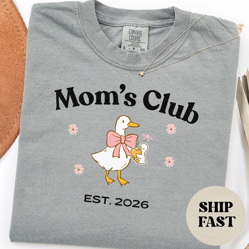 May include: A grey t-shirt with the words "Mom's Club" in black. The shirt features a cartoon duck wearing a pink bow and holding a baby duck. The text "EST. 2026" is below the graphic. A gold necklace is draped across the shirt.
