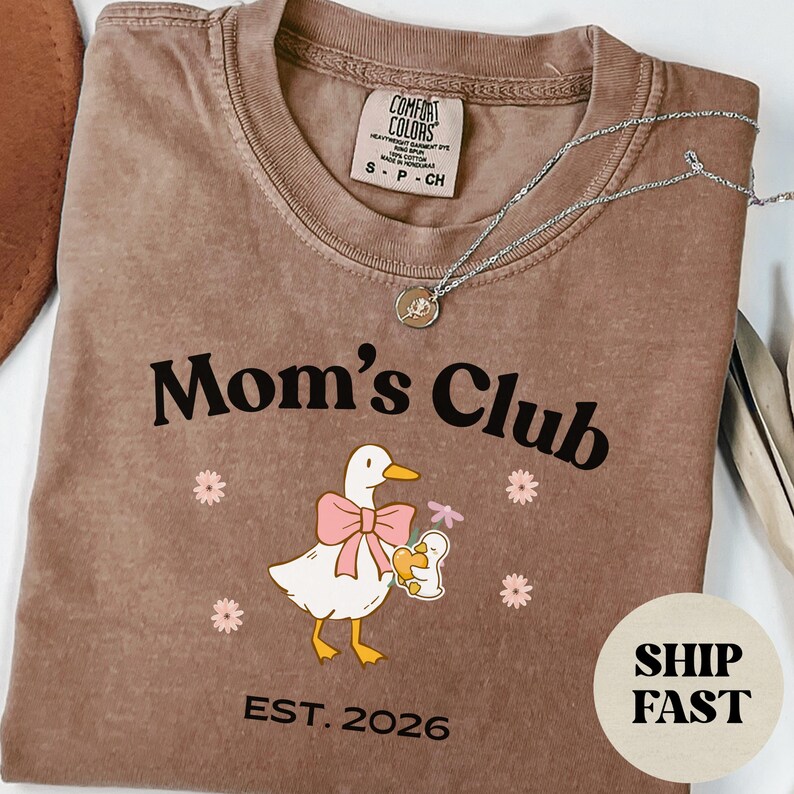 May include: A brown Comfort Colors t-shirt with the words "Mom's Club" and a duck graphic wearing a pink bow, holding a baby duck. The shirt also has "EST. 2026". A silver necklace with a small pendant is on the shirt.