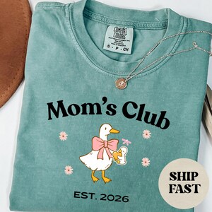 May include: A teal Comfort Colors t-shirt with the words "Mom's Club" in black. A cartoon duck with a pink bow and a baby duck graphic is below the text. The shirt also has the text "EST. 2026". A silver necklace with a pendant is on the shirt.