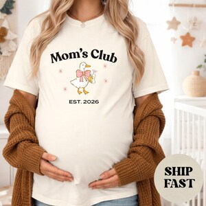 May include: A cream-coloured t-shirt with the words "Mom's Club" in black script. Below the text is a cartoon duck with a pink bow and flowers, and "EST. 2026" underneath. The shirt is worn by someone with long blonde hair.