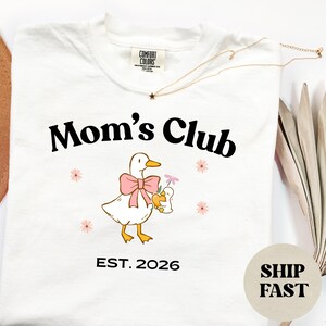 May include: Cream-coloured t-shirt with the words "Mom's Club" in black, a cartoon duck with a pink bow and a baby duck. The shirt also has the text "EST. 2026" and small pink flower graphics.
