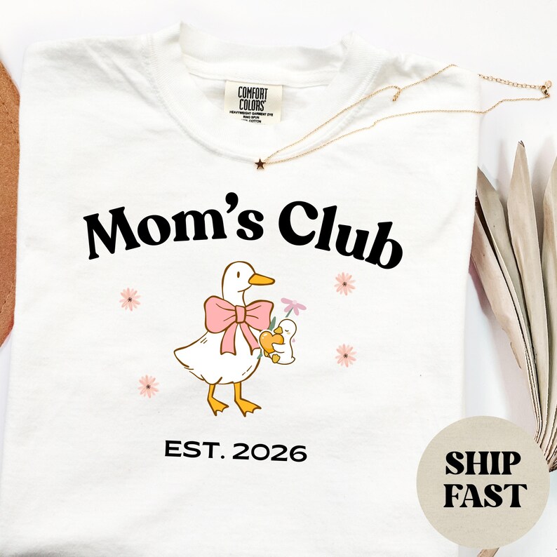 May include: Cream-coloured t-shirt with the words "Mom's Club" in black, a cartoon duck with a pink bow and a baby duck. The shirt also has the text "EST. 2026" and small pink flower graphics.