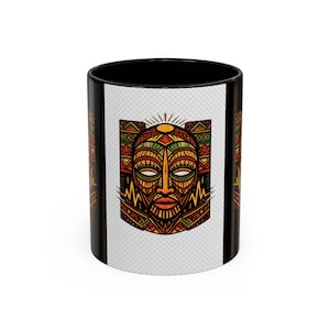 May include: Black coffee mug featuring a colorful tribal mask design. The mask is depicted with intricate patterns in shades of orange, yellow, green, and brown, set against a white background. The mug's interior and handle are black.