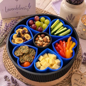 Könnte beinhalten: Ein schwarzes, rundes Snack-Tablett mit sechs blauen, herzförmigen Einsätzen und einem runden Behälter in der Mitte. Die Fächer sind mit Weintrauben, Gurkensticks, Nüssen, Paprikastreifen, Käsewürfeln und kernigen Crackern gefüllt.