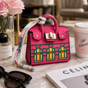 May include: A small, bright pink handbag with a textured surface, featuring a gold-tone metal turn-lock closure and a decorative light blue patterned silk scarf tied to the handle. The bag displays a green and yellow arched window graphic.
