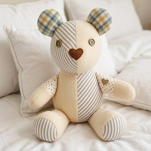 Pode incluir: Um urso de peluche feito de painéis de tecido patchwork. O urso apresenta lã de cor creme, secções às riscas cinzentas e brancas, orelhas aos quadrados e mangas com pintas. Tem dois olhos de botão castanhos e um nariz castanho em forma de coração.