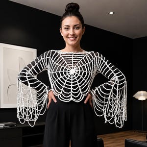 May include: A white long-sleeved crochet top featuring a prominent spider web pattern across the chest and back, with intricate dangling fringe details extending from the wide sleeves, worn over a black base layer with black high-waisted trousers.