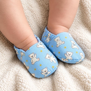 May include: A pair of light blue baby shoes featuring a repeating pattern of white teddy bears with small blue bows and tiny yellow stars. The slip-on footwear has an elasticated opening and a soft fabric construction for a comfortable fit.