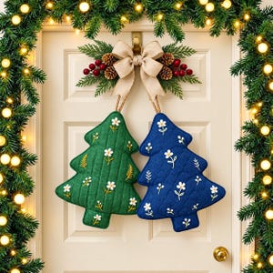 May include: Two quilted tree-shaped ornaments, one green and one blue, both featuring embroidered white flower patterns. They are suspended from a central beige fabric bow adorned with pine branches, pinecones, and red berries.
