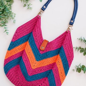 May include: A crochet hobo bag featuring a vibrant chevron pattern in magenta, teal, and orange. The bag includes a detachable dark blue leather handle attached with gold-tone metal clasps and a brown leather tab closure with a metal snap button.